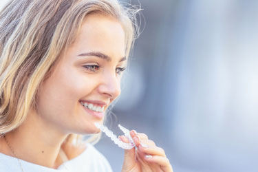Smiling blond woman in white shirt putting Invisalign in mouth